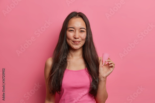 Positive dark haired Asian woman holds menstruation cup made of silicone or latex rubber as eco friendly alternative to pads and tampons, used during periods to prevent leaks. Gynecology, menses photo