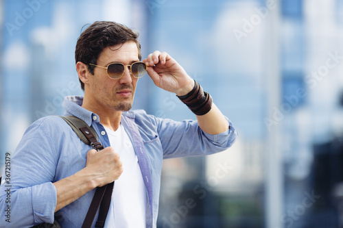 Rough unshaved handsom stylish male with backpack touches sunglasses