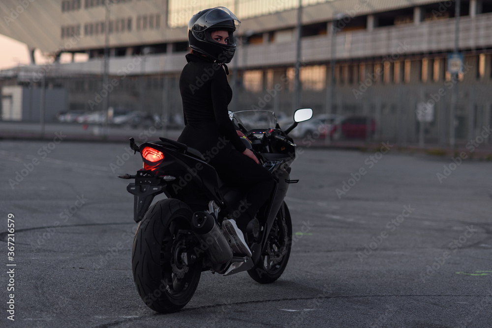 Adorable girl in black tight body suit and full-face helmet rides on stylish motorcycle at urban outdoors parking in evening. Freedom and active lifestyle concept. Totally black.