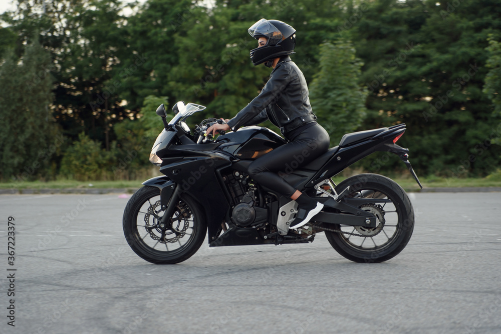 Stylish woman in black leather jacket, pants and full-face protective helmet rides on sports motorcycle at urban outdoors parking.