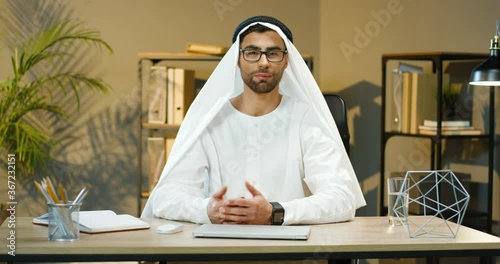 Young handsome muslim man in Kandura sitting at table in office and talking to camera while educating. Arabian business blogger or cauch in traditional emirates manswear speaking. Male of UAE. photo