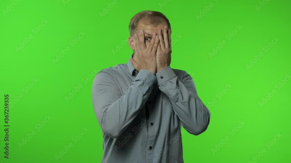 Shy guy scared to watch. Man covering eyes with hands and looking cautiously curious at camera