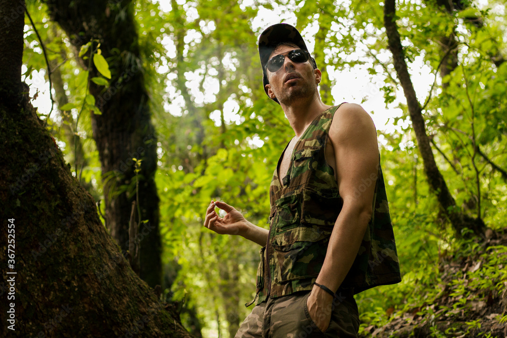 Guerrilla. Portrait of a young caucasian adult with strong arms wearing a camouflaged uniform and sunglasses, taking a smoke in the woods. 