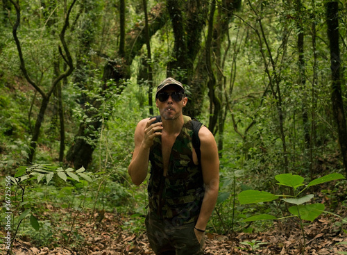 Guerrilla. Portrait of a young caucasian adult with camouflaged cloths in the jungle. Muscular man hiking with sunglasses and having a smoke.  © Gonzalo