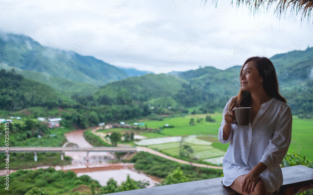 Portrait image of a beautiful asian woman holding and drinking hot coffee , sitting on balcony and looking at mountains and green nature