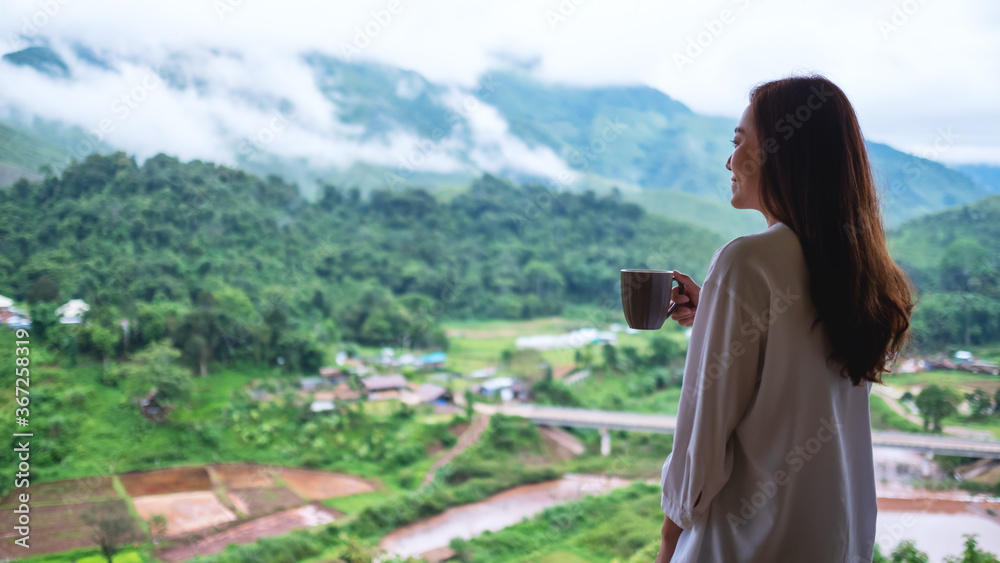 A beautiful asian woman holding and drinking hot coffee while looking at mountains and green nature