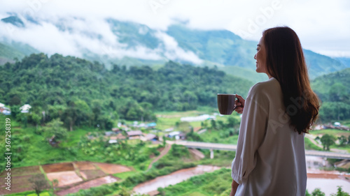 A beautiful asian woman holding and drinking hot coffee while looking at mountains and green nature © Farknot Architect