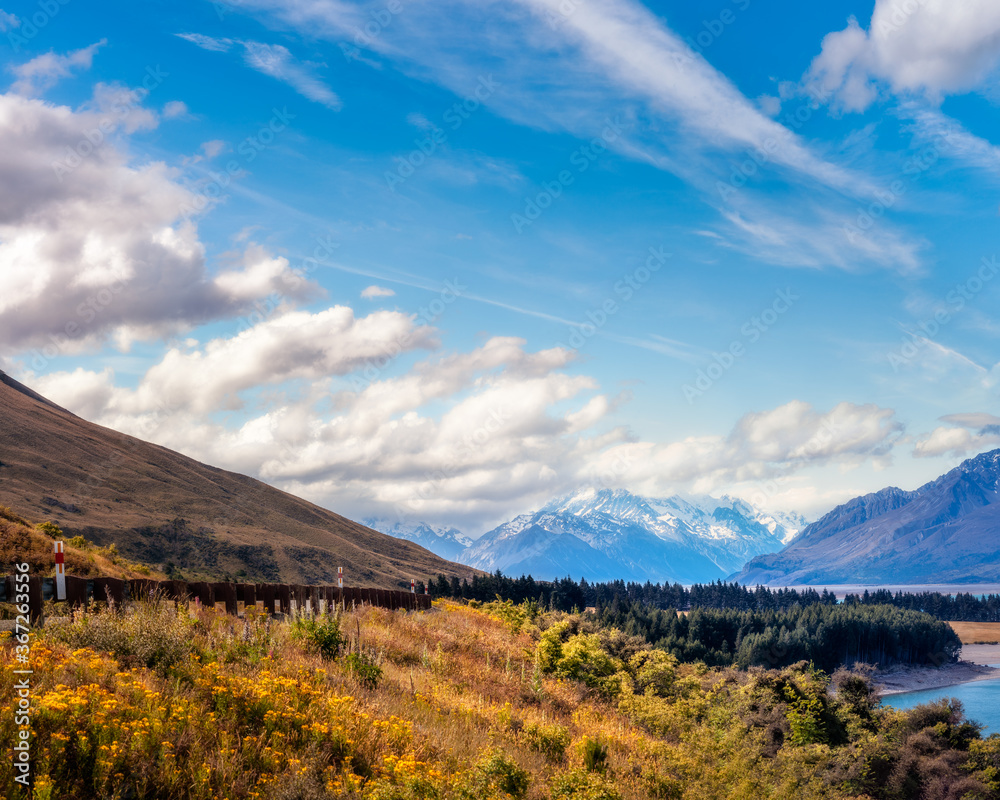Mount Cook Highway along the shore of Lake Pukaki, in New Zealand, South Island on a beautiful summer day with snow-capped Mount Cook engulfed in clouds in the background.