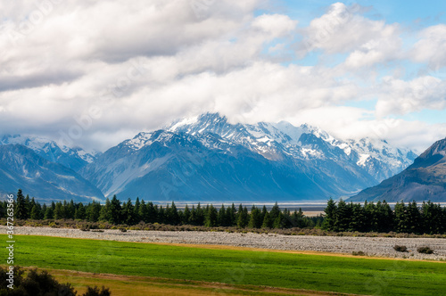 Close-up view of snow-capped Mount Cook engulfed in clouds, on the shore of Lake Pukaki, in New Zealand, South Island, with coniferous trees and green grass in the foreground.