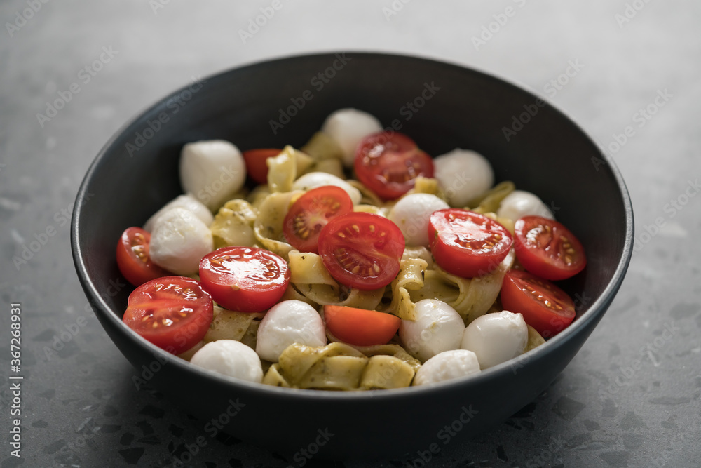 fettuccine pasta with pesto, mozzarella and cherry tomatoes in black bowl on concrete background fettuccine pasta with pesto, mozzarella and cherry tomatoes in black bowl on concrete background