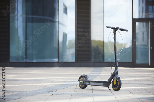 Background image of black electric scooter standing on tiled floor against glass building in urban setting, copy space