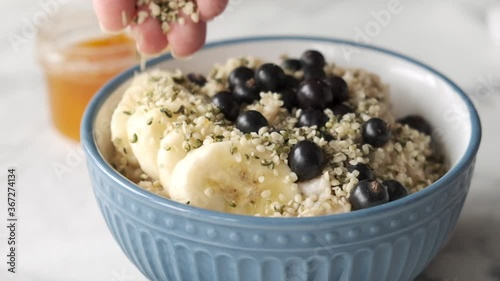 Slow motion adding hemp seeds into oatmeal porridge bowl with bananasand black currants. Healthy breakfast, vegan food concept