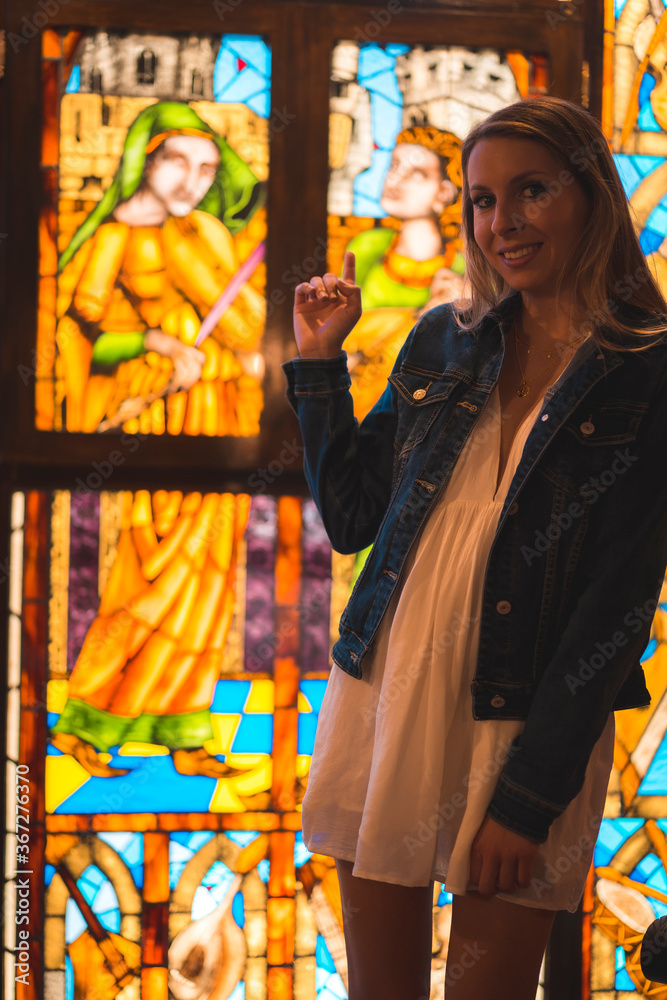 Young blonde caucasian woman in a white dress and denim jacket enjoying a beautiful medieval hotel in the town of Olite in Navarra. Spain, rural lifestyle. In a stained glass window