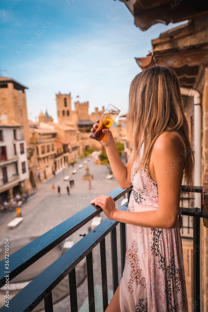 Young blonde caucasian woman in a long pink dress enjoying a beautiful rural hotel in Olite in Navarra. Spain, rural lifestyle. On the terrace with a soft drink and a castle in the background