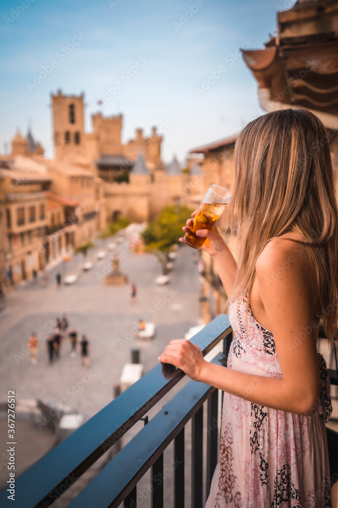 Young blonde caucasian woman in a long pink dress enjoying a beautiful rural hotel in Olite in Navarra. Spain, rural lifestyle. With a soft drink on the terrace and a castle in the background