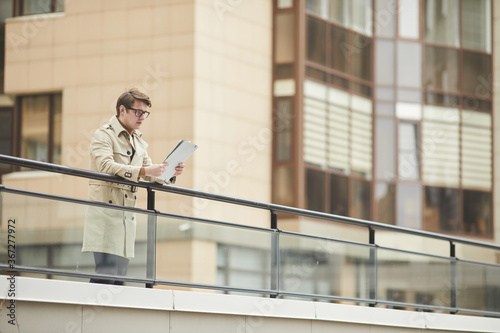 Wide angle portrait of modern young businessman wearing trenchcoat and reading document while leaning on railing outdoors in urban city setting, copy space