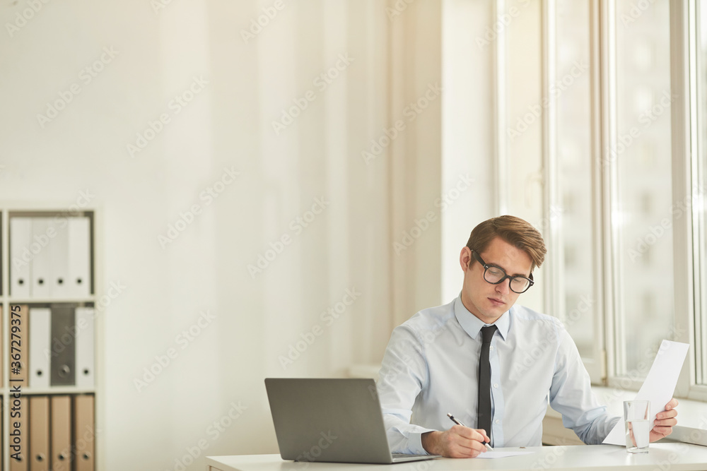 Minimal portrait of young successful businessman wearing glasses and using laptop while working at desk against white wall in office, copy space Minimal portrait of young successful businessman wearing glasses and using laptop while working at desk against white wall in office, copy space