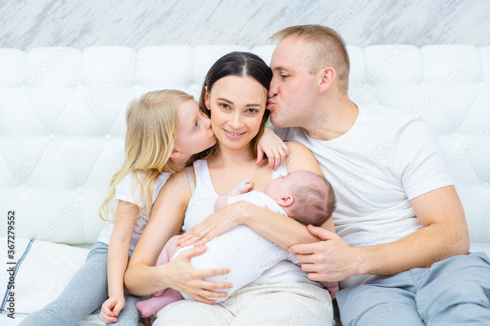 A newborn boy lies in his mother’s arms, a newborn hugs the mother of the newborn and the baby himself, and the older sister sits next to the mother. Happy family concept