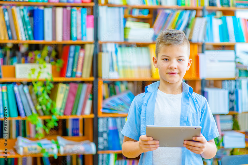 A boy in the library stands against the background of shelves with books and holds a tablet computer in his hands. Education and electronic systems concept A boy in the library stands against the background of shelves with books and holds a tablet computer in his hands. Education and electronic systems concept