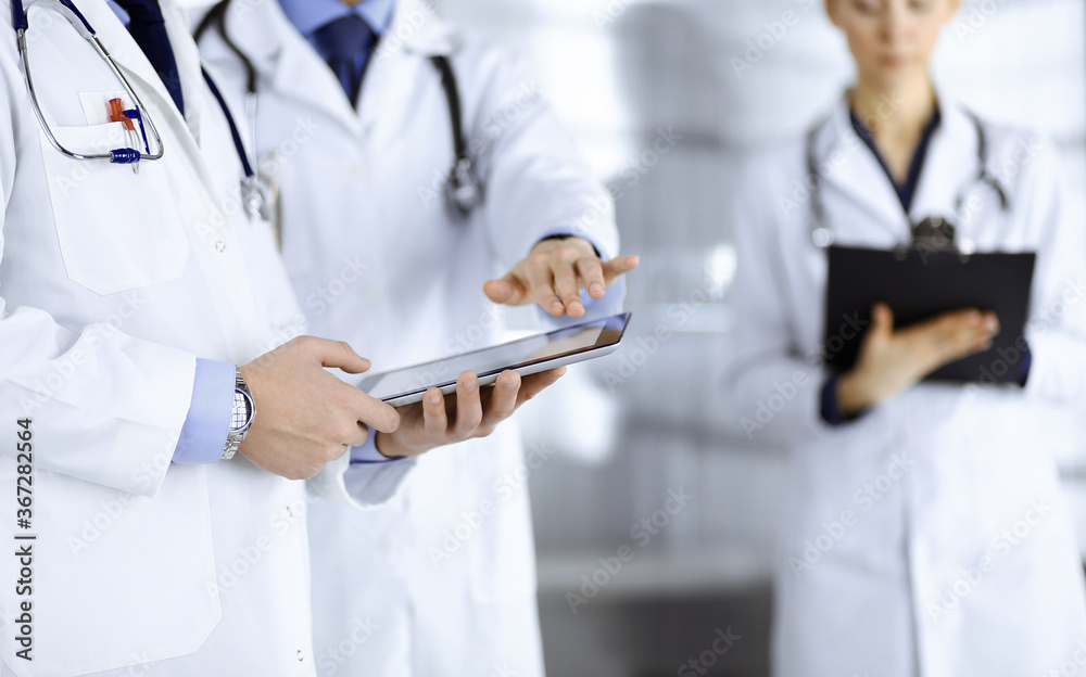 Group of unknown doctors use a computer tablet to check up some medical names records, while standing in a hospital office. Physicians ready to examine and help patients. Medical help, insurance in