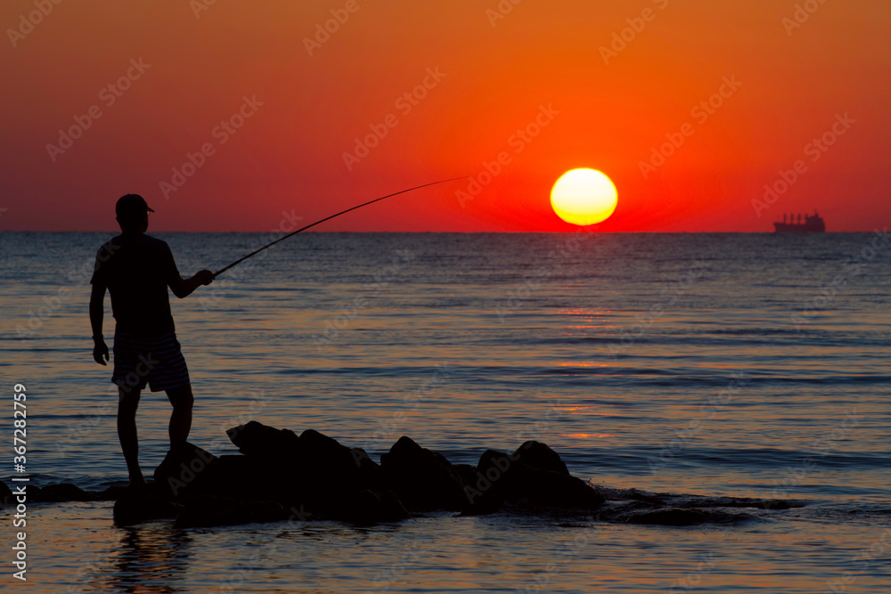 A fisherman catches fish on the seashore in the early summer morning. Silhouette of a fisherman on the background of the rising sun. Selective focus.