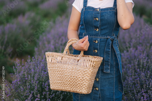 Middle selection of Attractive young woman walking in summer lavender field. Model dressed in denim sundress with straw hat and bag. Close up