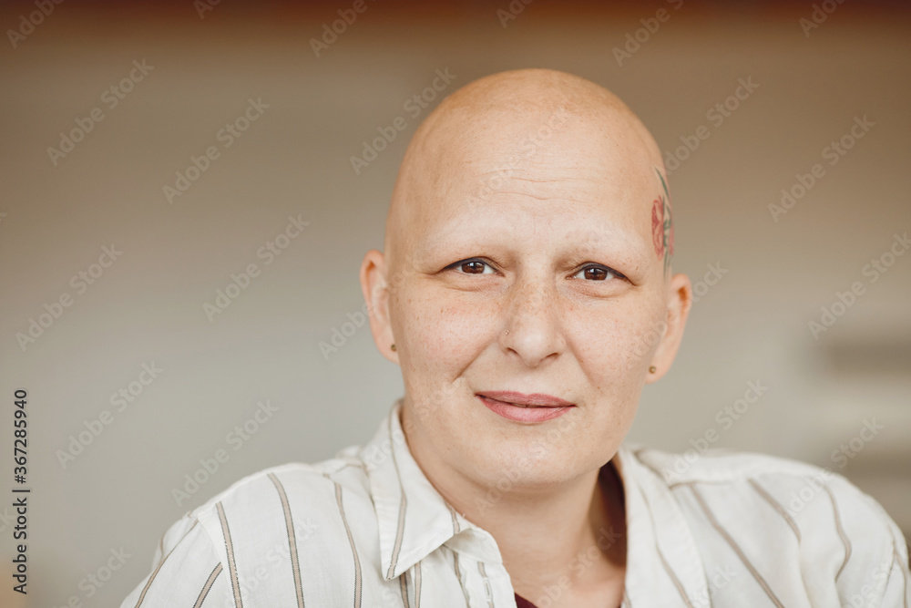 Fototapeta premium Minimal head and shoulders portrait of bald adult woman looking at camera and smiling while sitting on couch in warm-toned interior, alopecia and cancer awareness, copy space