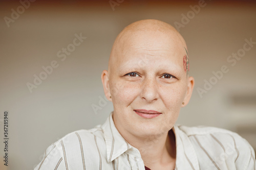 Minimal head and shoulders portrait of bald adult woman looking at camera and smiling while sitting on couch in warm-toned interior, alopecia and cancer awareness, copy space photo