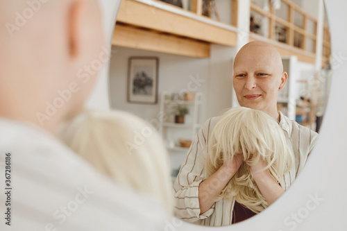 Reflection portrait of bald adult woman looking in mirror holding wig while standing in warm-toned home interior, alopecia and cancer awareness, copy space photo