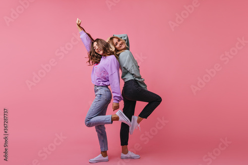 Full-length photo of inspired girls funny dancing together. Indoor shot of cheerful best friends standing on bright background.