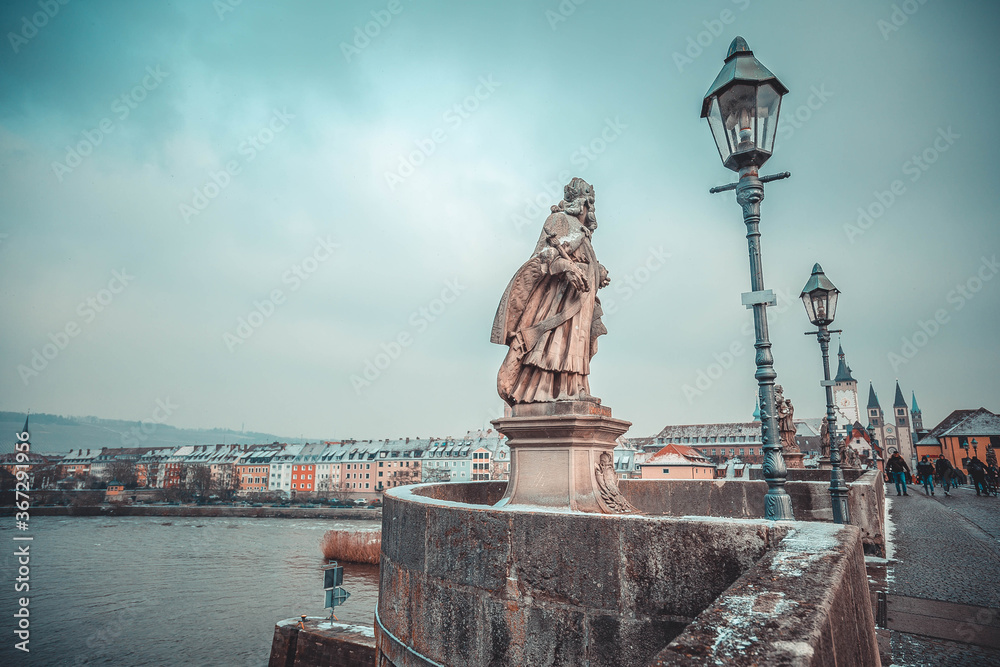 Fototapeta premium snowfall on the Main bridge of Wuerzburg, Frankonia, Bavaria, Germany. Snow covered statues, lamp posts, houses, buildings, churches. Foot marks on snow. Hazy morning. Main river. Snow on the cliff. 