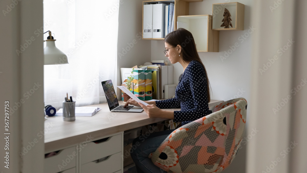 Focused millennial Caucasian woman in glasses sit at desk at home work on laptop with documents, concentrated female or girl study on computer, prepare report research, distant education concept Focused millennial Caucasian woman in glasses sit at desk at home work on laptop with documents, concentrated female or girl study on computer, prepare report research, distant education concept
