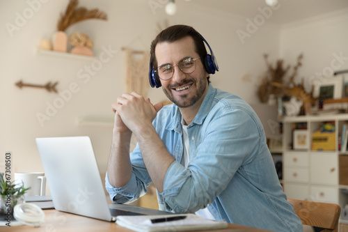 Portrait of smiling millennial male coach in headphones sit at desk at home working on laptop, happy motivated young Caucasian man in headset and glasses study distant watch webinar on computer