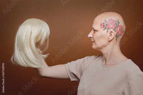 Fototapeta Side view portrait of confident bald woman holding wig of blonde hair against br