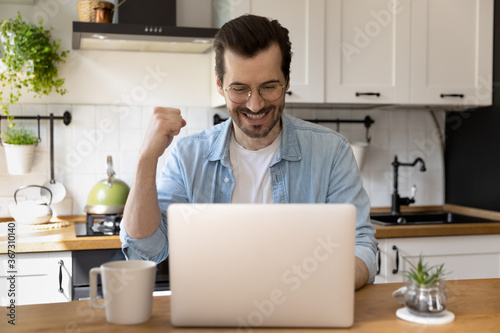 Overjoyed young Caucasian man in glasses sit at desk in kitchen look at laptop screen read good promotion news online, happy millennial male triumph excited with internet lottery win on computer