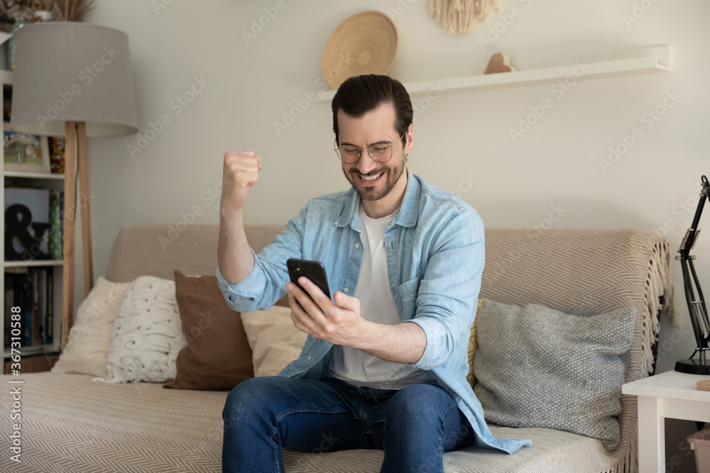 Excited millennial Caucasian male sit on couch at home triumph get pleasant good message on cellphone, overjoyed young man feel euphoric with online lottery win victory on smartphone, success concept Excited millennial Caucasian male sit on couch at home triumph get pleasant good message on cellphone, overjoyed young man feel euphoric with online lottery win victory on smartphone, success concept