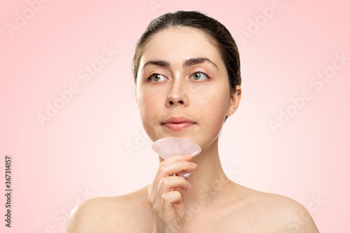 Gua Sha Massage. A woman in a white bath towel massages her chin with a quartz guasha scraper. Pink background. Copy space. Concept of alternative medicine and cosmetology