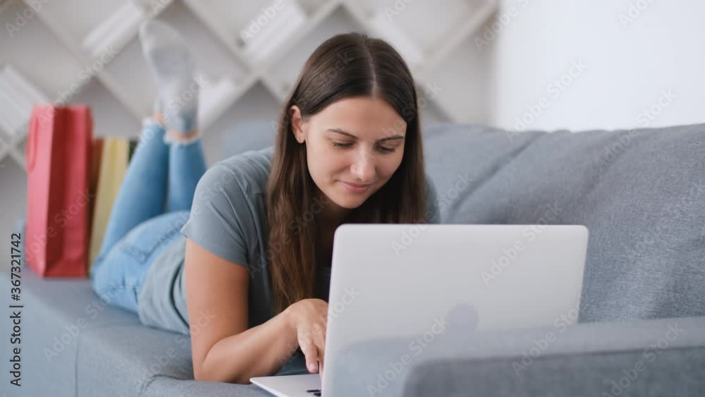Online shopping, Buying from home, Credit card, Online store. Woman lying on the couch and choosing a product from an online store for home delivery using a laptop Online shopping, Buying from home, Credit card, Online store. Woman lying on the couch and choosing a product from an online store for home delivery using a laptop