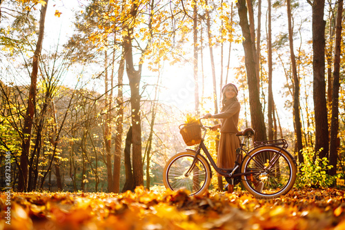 Fototapeta Naklejka Na Ścianę i Meble -  Young smiling girl with a bicycle walks in the autumn forest at sunset.