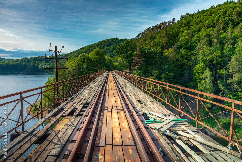 Obraz premium Historic steel truss underspanned suspension railway bridge from 1902 over Bobr River in Pilchowice, Poland at beautiful morning. Former German engineering construction at closed railway line