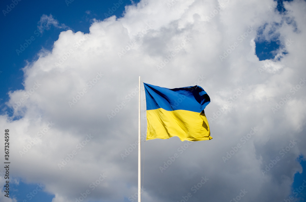 Blue and yellow Ukrainian national flag on a flagpole against a blue sky and white clouds, flag waving in the wind Blue and yellow Ukrainian national flag on a flagpole against a blue sky and white clouds, flag waving in the wind
