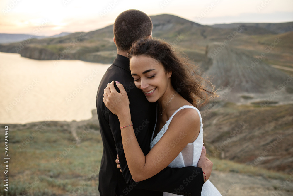 Happy bride gently embraces her beloved. Beautiful bride closes her eyes and smiles in the arms of a spouse. Lovers stand against the background of the bay and hills at sunset. Happy bride gently embraces her beloved. Beautiful bride closes her eyes and smiles in the arms of a spouse. Lovers stand against the background of the bay and hills at sunset.