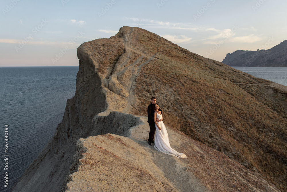 Wedding couple standing on a cliff against the background of the sea and mountains. A groom gently embraces his beautiful bride in fashion wedding dress with long hem.
