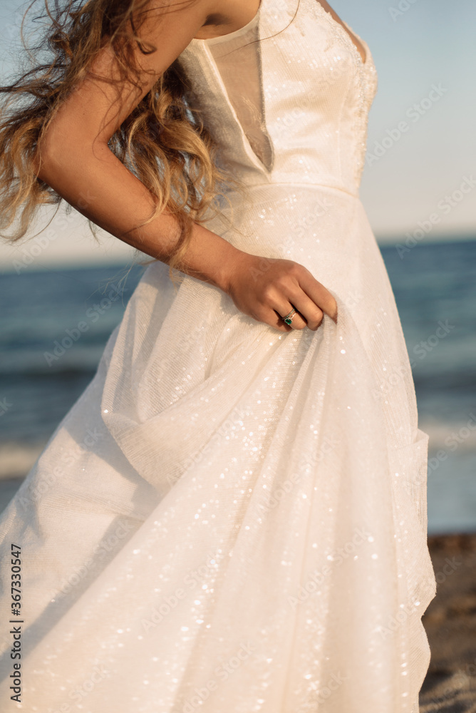 Close-up photo of luxury wedding dress details. Bride with beautiful engagement green ring on her finger holds her sparkling dress on the beach. Close-up photo of luxury wedding dress details. Bride with beautiful engagement green ring on her finger holds her sparkling dress on the beach.