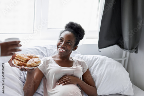 kind african american man takes care of his pregnant wife, he gives breakfast to her, woman lie on bed in the morning, happy relationships