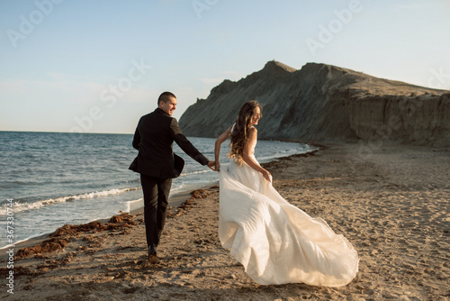 Fotografie Beautiful wedding couple run along the beach near the coastline holding hands against the background of rocks in the sea