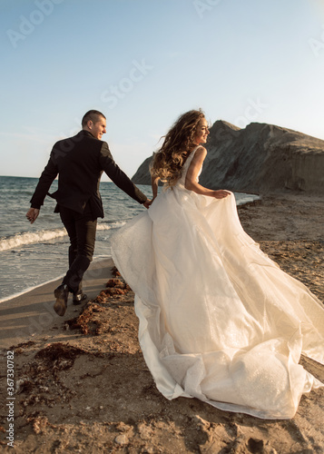 Foto Happy wedding couple run along the beach near the coastline holding hands against the background of rocks in the sea