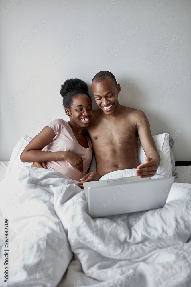 happy black married couple have rest on bed in the morning, african american woman sits with laptop, man lie on bed next to her.