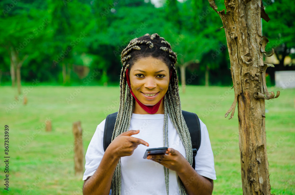 Naklejka premium young beautiful african student wearing face mask preventing herself from the outbreak in the society feeling excited about what she saw on her cellphone and points to her phone