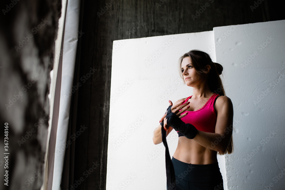 Obraz premium Portrait of a young woman kickboxer in a pink top who ties a black elastic bandage on her hand before fighting and training on a white background.